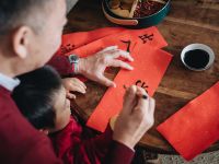 Grandfather Practising Chinese Calligraphy For Chinese New Year Fai Chun (Auspicious Messages) And Teaching His Grandson By Writing On Couplets At Home